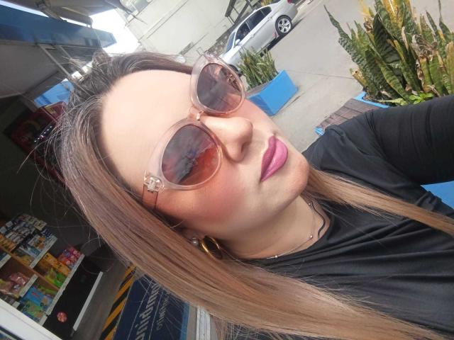 AvaMia, com óculos de sol com tons de rosa, batom rosa e cabelos castanhos longos, vestindo uma camisa preta e brincos de argola dourados, tirando uma selfie ao ar livre..