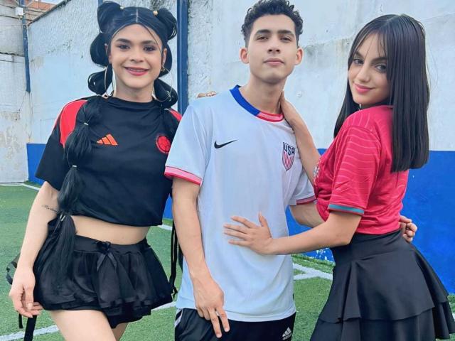 Drei Personen stehen da. DreamsWetHot trägt einen schwarzen Sport-BH und einen Rock, hat kleine Brüste. DreamsWetHot trägt ein weißes Fußballtrikot. DreamsWetHot trägt ein pinkes Hemd und einen schwarzen Rock. Alle lächeln..