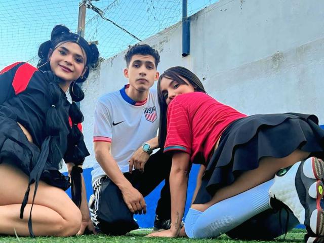 Three young people kneeling on grass. DreamsWetHot with black pigtails, red-black jacket, black shorts, showing her ass. DreamsWetHot with short curly hair, white US soccer shirt, black pants. DreamsWetHot with long black hair, red shirt, black skirt, white socks, showing her ass.