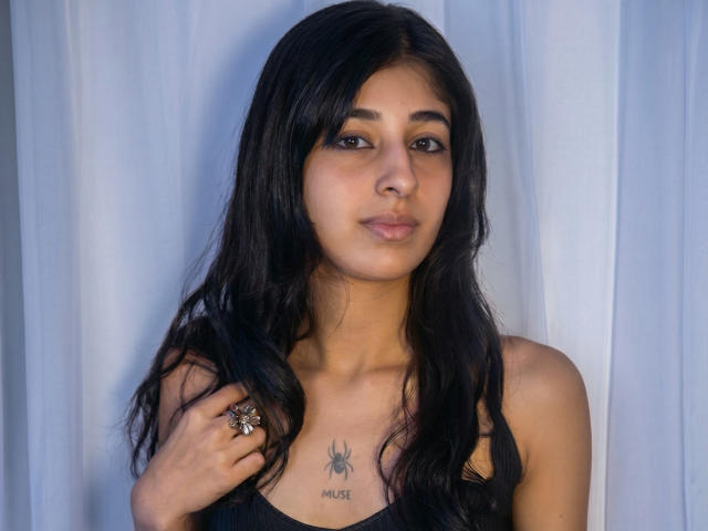 KeylaStone has dark, wavy hair, brown eyes, and a spider tattoo on her chest. She's wearing a black top, holding her hair, and has a ring on her right hand.