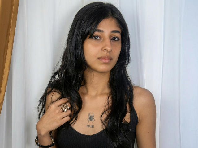 KeylaStone with long black hair, brown eyes, tan skin, small tits, black tank top, spider tattoo on chest, holding ring, white curtain behind, golden curtain to the left.