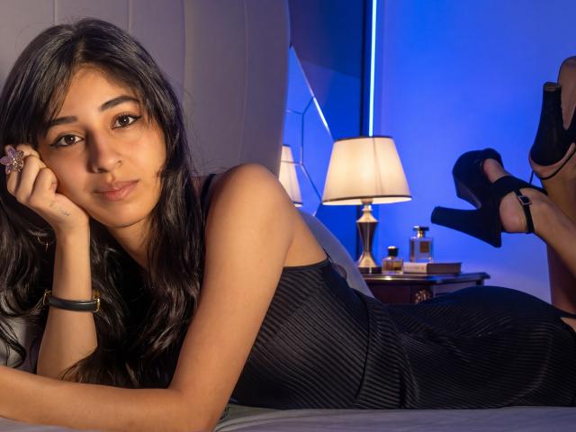 KeylaStone lying on a bed in a black, ribbed dress, looking at the camera with a slight pout, her black heels in the air, blue light in the background.