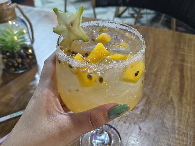 MarilynHills' hand with green nails holds a yellow drink with ice, pineapple chunks, blackberries, and a star anise on a salted rim, on a wooden table.