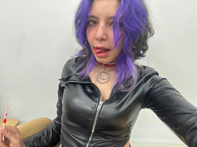 DarckVelvet with purple hair, black leather zip-up, red choker, and ring, holding candy, winking, and parting lips, showing tits, in a selfie.