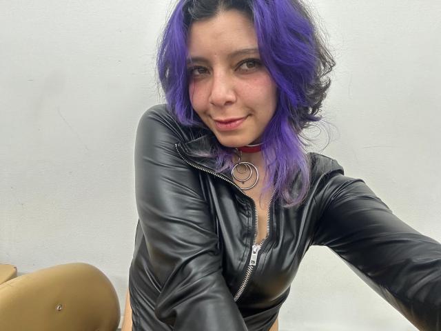 DarckVelvet with purple hair, black leather zip-up, and red choker, smiling at camera, showing a cute face with slight blush. Her tits are small, she's sexy.