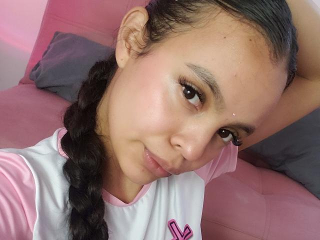 DanesaCronin with a braid, pink and white shirt, small nose piercing, brown eyes, subtle smile, pinkish skin, looking at camera, hand in hair, pink and gray background.
