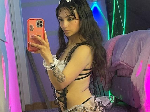 MarietteJones with long black hair, wearing a black lace bra and white scrunchie, holding a pink phone, showing tattoos, looking in a mirror, showing her small tits and ass.
