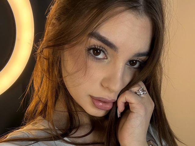 RoseleeVandiest with brown eyes, full pink lips, and long brown hair, touching her cheek with a ringed hand, looking directly at the camera, wearing a gray top.