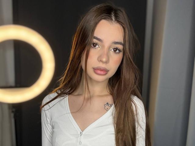 Roselee Vandiest a de longs cheveux bruns, des yeux bruns et un petit tatouage sur sa clavicule droite. Elle porte une chemise blanche à fermeture éclair et regarde directement l'appareil photo..