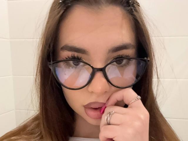 RoseleeVandiest with glasses, brown hair, and pink lipstick, touching her lips with a ringed finger, looking seductively at the camera, her reflection in the glasses is visible.