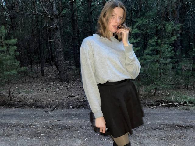 JennellMatacale, with blonde hair, gray sweater, black skirt, and tan tights, stands in a forest, touching her lips with a finger, showing a subtle smile.