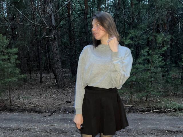JennellMatacale in a gray sweater, black skirt, and brown hair, standing in a forest, looking to the side with a hand near her face. Her ass is small.