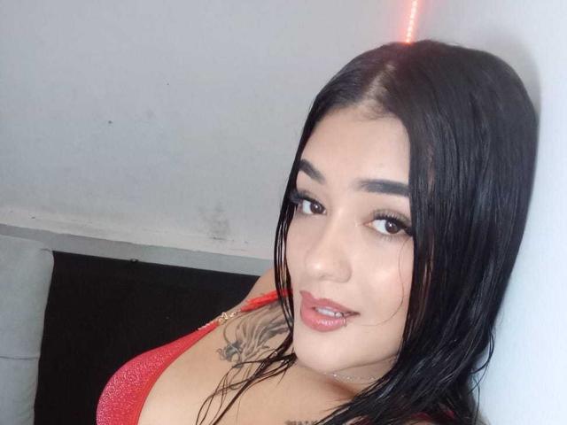 BrianaXO mit schwarzen Haaren, rotem Lippenstift und einem Tattoo an der rechten Schulter, trägt einen roten BH und blickt mit einem leichten Lächeln in die Kamera..