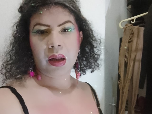 MistressEmeraMt has curly black hair, green and yellow eyeshadow, pink lipstick, and white makeup on her face. She wears pink earrings and a black top, looking surprised.