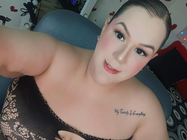GiiannaMorrys with her tits showing in a lace black top, looking at the camera with a smile, short brown hair, tattoo on her right shoulder, fair skin, and red lipstick.