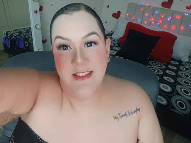 GiiannaMorrys with dark hair pulled back, fair skin, blue eyes, red lipstick, nose ring, tattoo on right shoulder, smiling, topless, showing large tits, in a bedroom with bed.