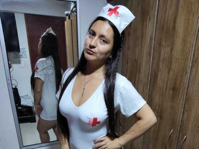 VenusSweetPalmer in a white nurse outfit with red crosses, long black hair, and a nurse hat, standing with hands on hips, reflecting in a mirror, showing her tits.