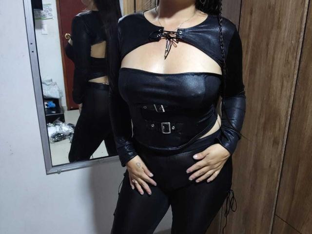 VenusSweetPalmer in black leather outfit, showing tits through cutout, lace-up top, belt, and tight pants, hands on hips, mirror reflection, wood cabinet, dark hair, bedroom.