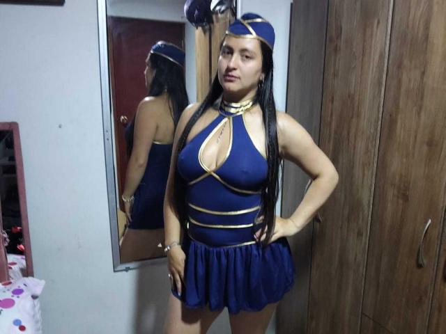 VenusSweetPalmer stands in a room, wearing a blue and gold costume with a cutout top, revealing her big boobs, and a short skirt, with a matching hat.