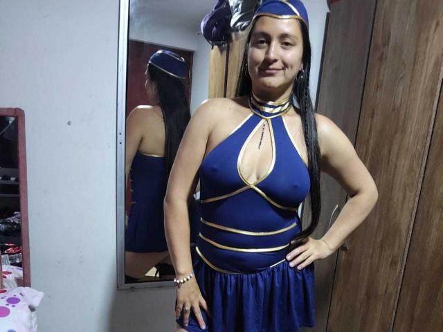 VenusSweetPalmer, wearing a blue and gold dress with a halter neck and cutout, has long black hair, a smile, and stands in front of a mirror, showing her tits.