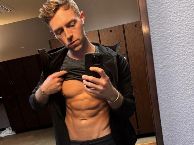 BlakeOnCam, with a fit, toned abs, pulls up his shirt in a mirror selfie, wearing a black jacket and grey tee, holding a phone, looking serious, short blonde hair.