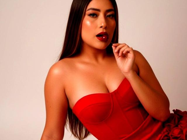 VelvetRoseX with long brown hair, red lipstick, and shiny skin, wearing a strapless red dress that highlights her tits, holding her finger to her mouth seductively.