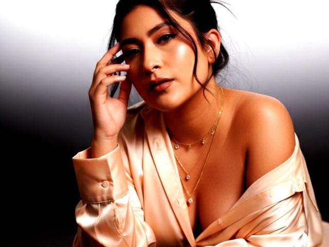 VelvetRoseX with shiny, off-shoulder, cream satin shirt, hand near face, gold necklace, and subtle makeup, showing her tits, dark hair in messy bun, confident, sexy look.