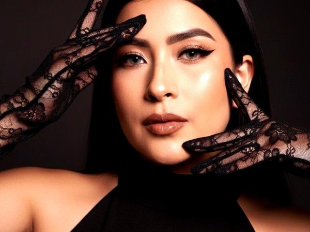 VelvetRoseX, con guantes de encaje negro, tocándose la cara, tiene una piel brillante, labios carnosos y cejas oscuras, y lleva un top de tirantes negro, mirando seductoramente a la cámara..