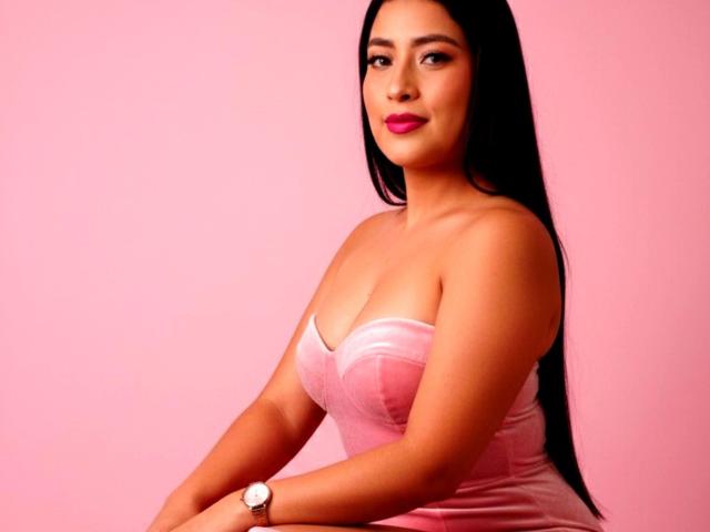 VelvetRoseX met lang zwart haar, een roze jurk zonder bandjes, roze lippenstift en een horloge om haar pols, zittend voor een roze achtergrond. Haar borsten zijn zichtbaar, ze kijkt iets opzij en heeft een zelfverzekerde glimlach..