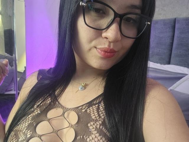 LunaBellaHotty with black hair, glasses, and a smile, wearing a revealing black fishnet top that shows her tits. She's got a small necklace and pink lipstick.