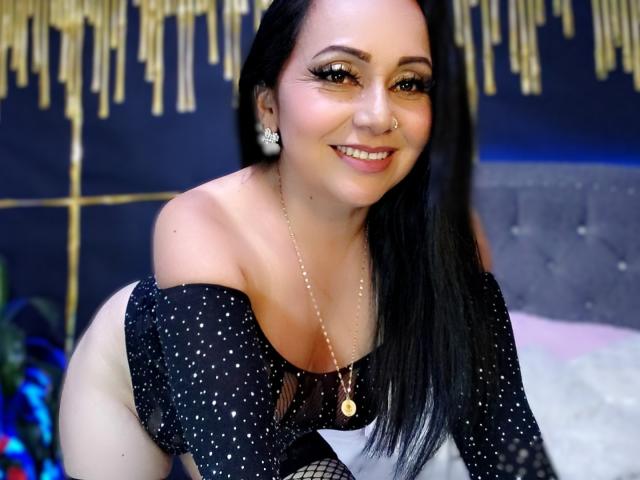 Zaria69 with black hair, smiling, wearing a black off-shoulder polka dot top, revealing her tits, nose ring, and necklace, leaning forward.