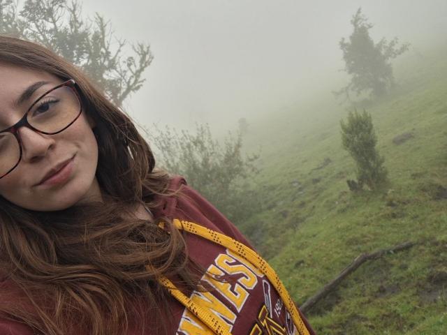 Victoria Palmer, con cabello castaño largo, gafas y una camisa color burdeos, con los senos ligeramente visibles debajo de una chaqueta amarilla, de pie en una colina verde y cubierta de niebla..