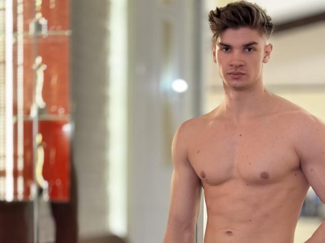 MaxKaiden, shirtless, with a toned, muscular, white chest and abs, standing in a room, brown hair, looking at the camera, neutral expression, slightly blurred background.