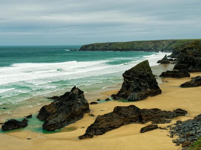 Una playa rocosa con arena color arena, rocas oscuras y puntiagudas, y olas verde azuladas. Los pechos de GlitterOneTest-hot son la espuma blanca del océano, y su trasero es el acantilado en el fondo..
