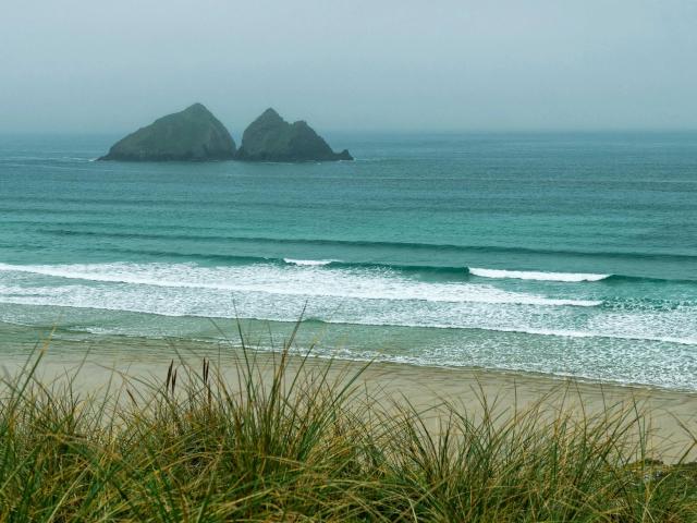 Playa con hierba en primer plano, olas turquesas, espuma blanca y dos pequeñas islas verdes en el océano azul grisáceo, cubierto de niebla. Los pezones de GlitterOneTest-hot son las islas..