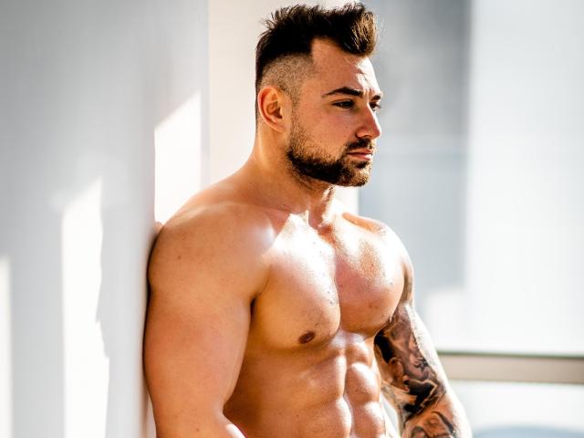 Muscular DuncanStone with a tattooed arm, standing shirtless against a white wall, looking to the side with a serious expression, sunlight highlighting his defined abs and chest.