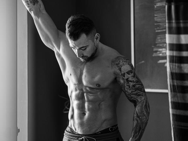 DuncanStone, shirtless, muscular, tattooed arm, standing by window, arm raised, looking down, Calvin underwear, defined abs, short beard, black and white, serious expression, window light.