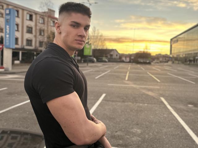 MaxKaiden, muscular, with short black hair, wearing a black t-shirt, standing in an empty parking lot, arms crossed, looking back over shoulder, sunset in background.