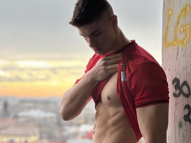 MaxKaiden with short dark hair, wearing a red shirt, exposing a toned, muscular chest, holding the shirt with his left hand, a silver pen on the shirt.