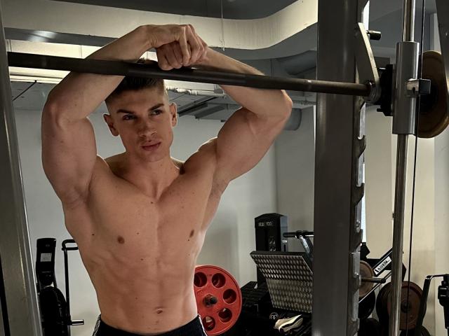 Muscular MaxKaiden with a chiseled upper body, gripping a bar, doing overhead workouts in a gym. His bare, taut chest and black shorts are visible.