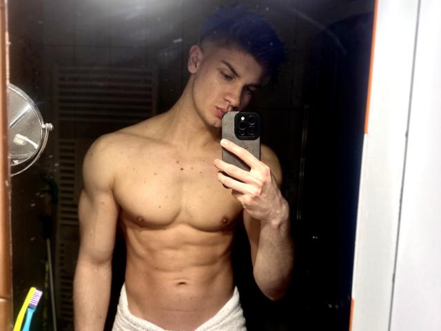 Mirror selfie of MaxKaiden, shirtless, showing a muscular, toned chest and abs, with a white towel around his ass, holding a black phone, in a dimly lit room.