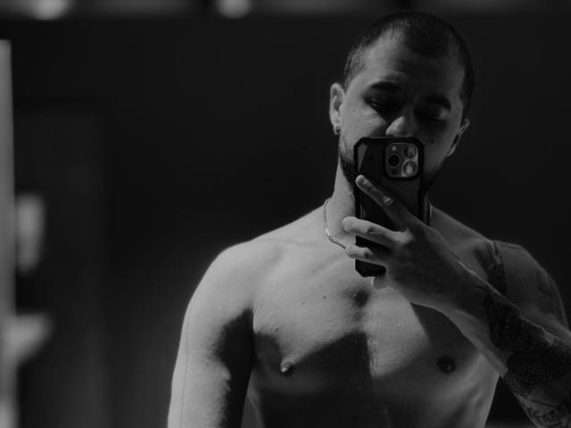 Shirtless JamesMyers with a tattooed arm, holding a phone to his face, looking in a mirror, dark room, blurred background, phone case with a circular pattern.