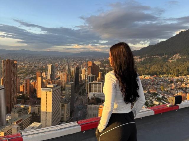 BerlyVenuss with long dark hair, white long-sleeve shirt, black pants, standing on rooftop, hands in pockets, looking at city skyline, mountains in background, sunset, cloudy sky.