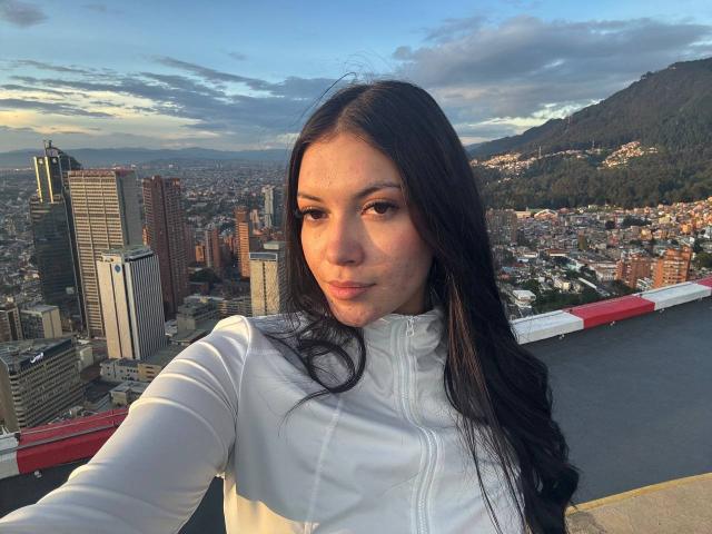 BerlyVenuss has long black hair, wearing a white long-sleeve shirt, standing on a rooftop with cityscape and mountains in the background, sunlight on her face, looking at camera.