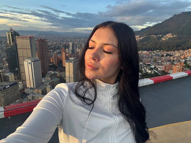 BerlyVenuss with closed eyes, long black hair, white long-sleeve shirt, slightly pouty lips, standing on rooftop, city and mountains in background, sunset lighting, calm expression.