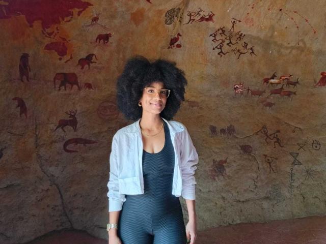 Viiolettaaa, con i capelli neri ricci, sorride, indossa una giacca bianca sopra una tuta nera con un motivo a zig-zag, ed è in piedi contro un muro con antiche pitture rupestri. Il seno è piccolo. Le natiche non sono visibili..