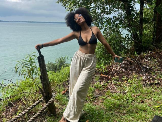 Viiolettaaa with dark curly hair, black bra, and beige pants, leaning on a rope by a lake, smiling, her tits slightly visible, standing on grassy, wooded terrain.