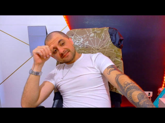 JamesMyers, bald with a tattooed arm, wearing a white tee, sits in a chair, holding his head, looking slightly annoyed, with a cracked wooden backrest behind him.
