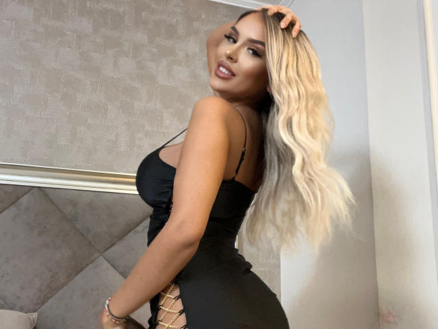SelenaVoss with long, wavy blonde hair, black dress, showing her ass, looking over her shoulder, hand in hair, smiling, wearing bracelets, in a bedroom.