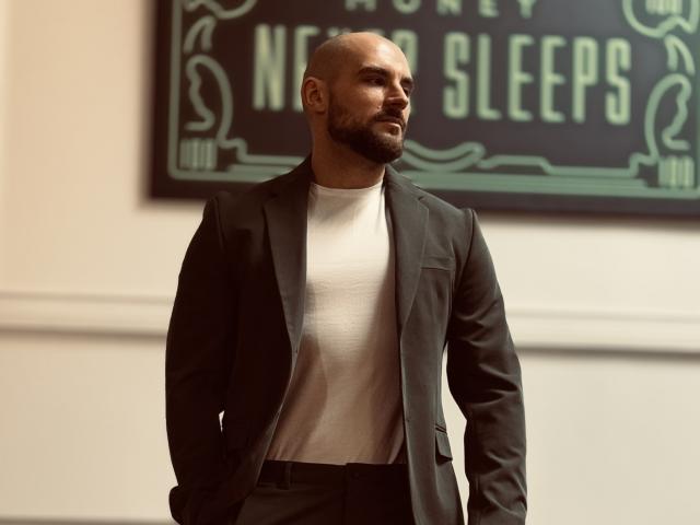 AdamJameson, bald with a beard, wearing a black suit over a white t-shirt, standing with hands in pockets, looking to the side, in a well-lit room.